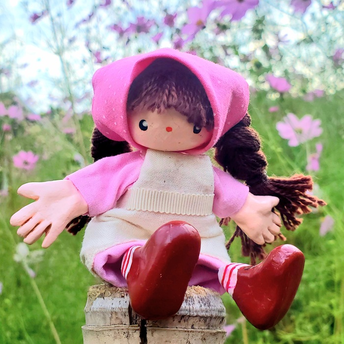 手作りの女の子のぬいぐるみ。ピンクの服と帽子を着て、木のベンチに座っている。背景は緑と花畑。